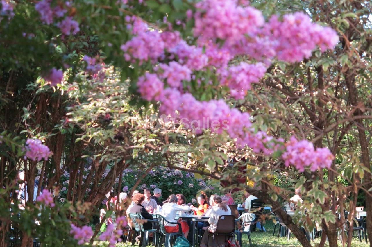 Des personnes assises à des tables dans un jardin, entourées de buissons fleuris aux fleurs roses.