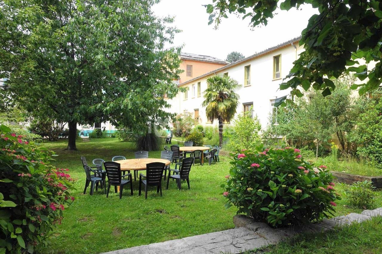 Jardin verdoyant avec tables et chaises en plastique noir, entouré d'arbres et de buissons fleuris devant un bâtiment clair.