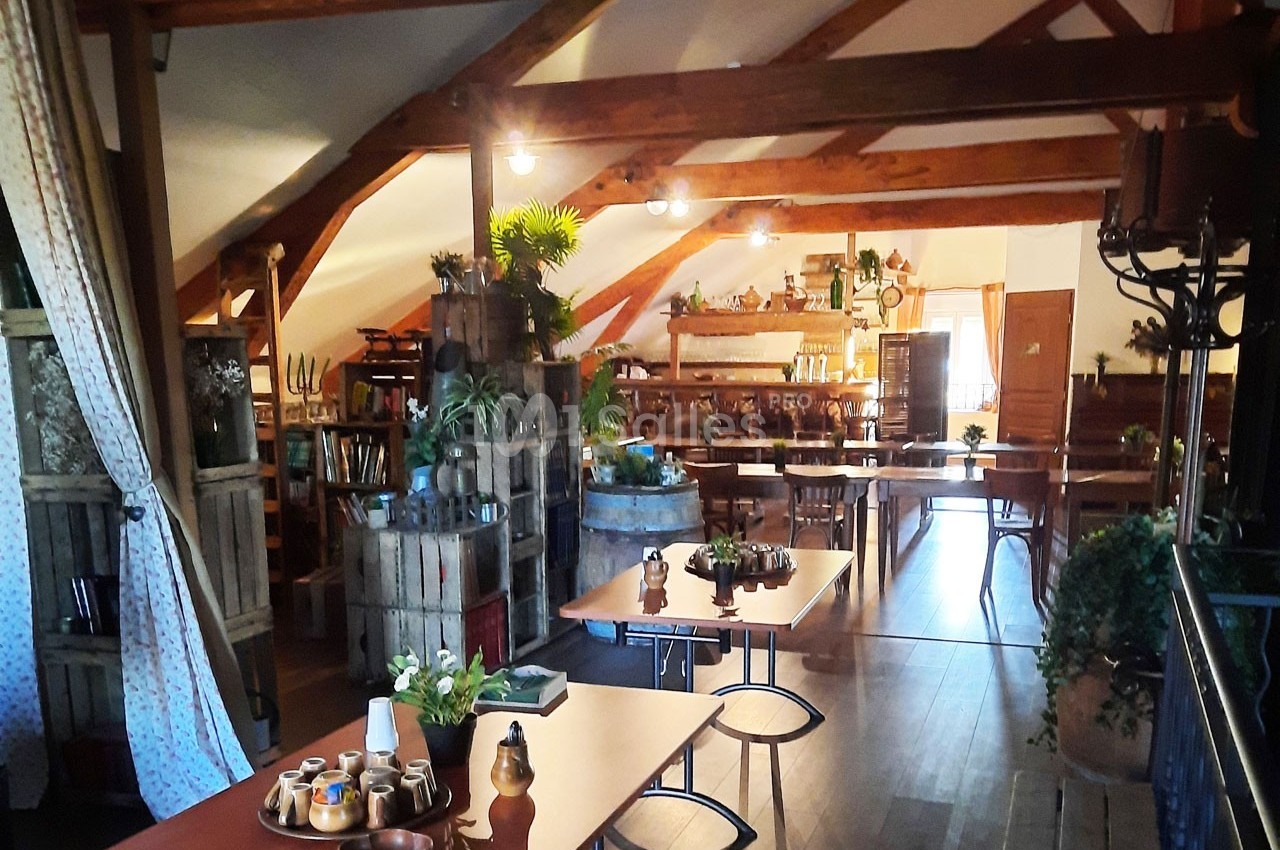 Salle lumineuse avec poutres apparentes, tables en bois, plantes décoratives et ambiance rustique chaleureuse.