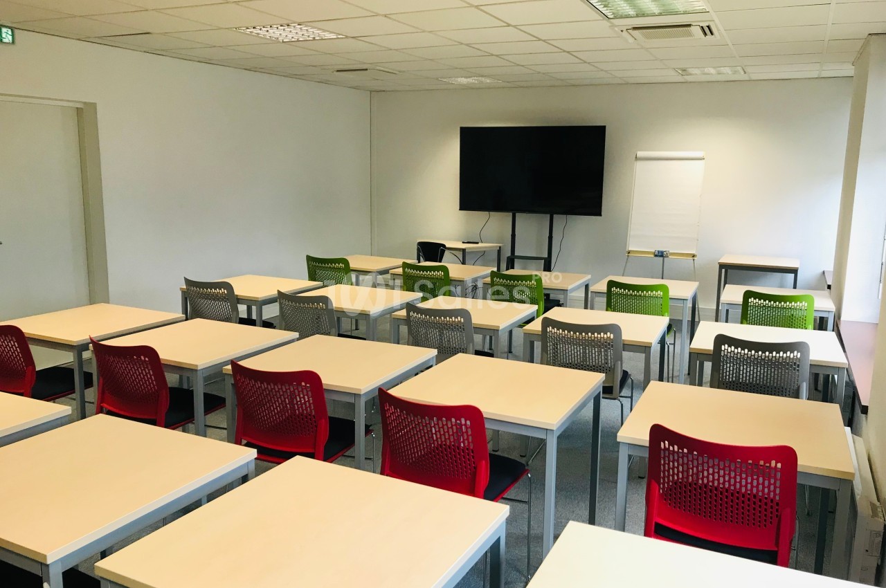 Salle de classe vide avec des tables, des chaises colorées, un tableau blanc, un écran et un paperboard.