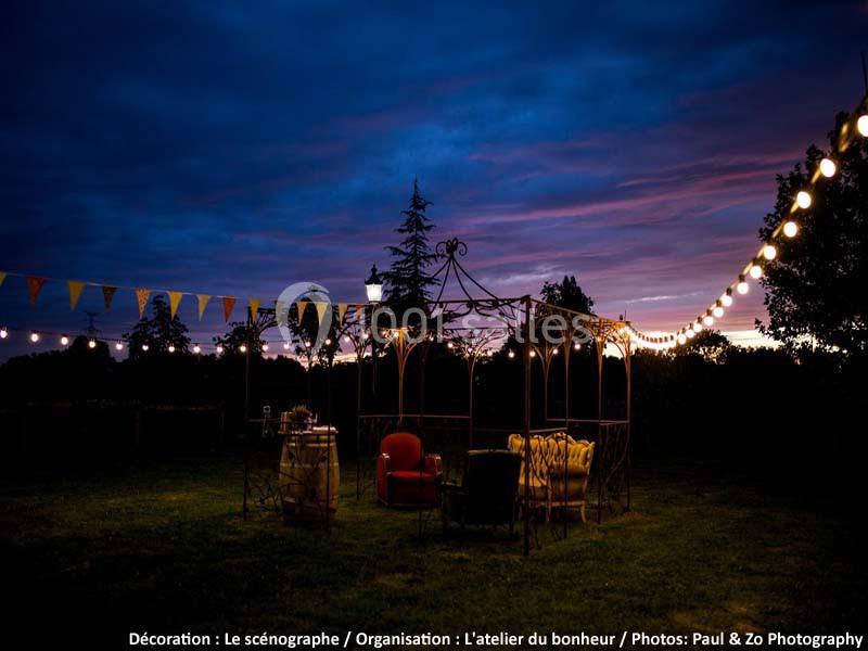Espace extérieur décoré avec guirlandes lumineuses, fauteuils et tonnelle, au crépuscule dans un jardin.
