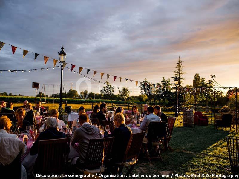 Des convives dînent en extérieur au coucher du soleil, entourés de guirlandes lumineuses et de fanions colorés.