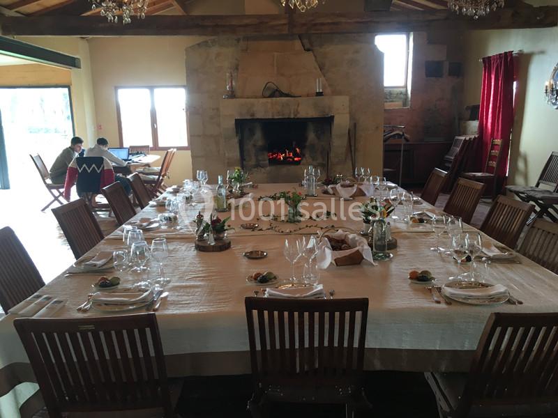 Grande table dressée avec vaisselle et décorations, dans une salle chaleureuse avec cheminée allumée et poutres apparentes.