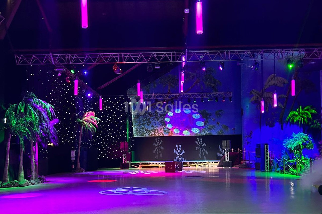 Salle de spectacle décorée avec des palmiers artificiels, éclairages colorés et projecteurs lumineux.