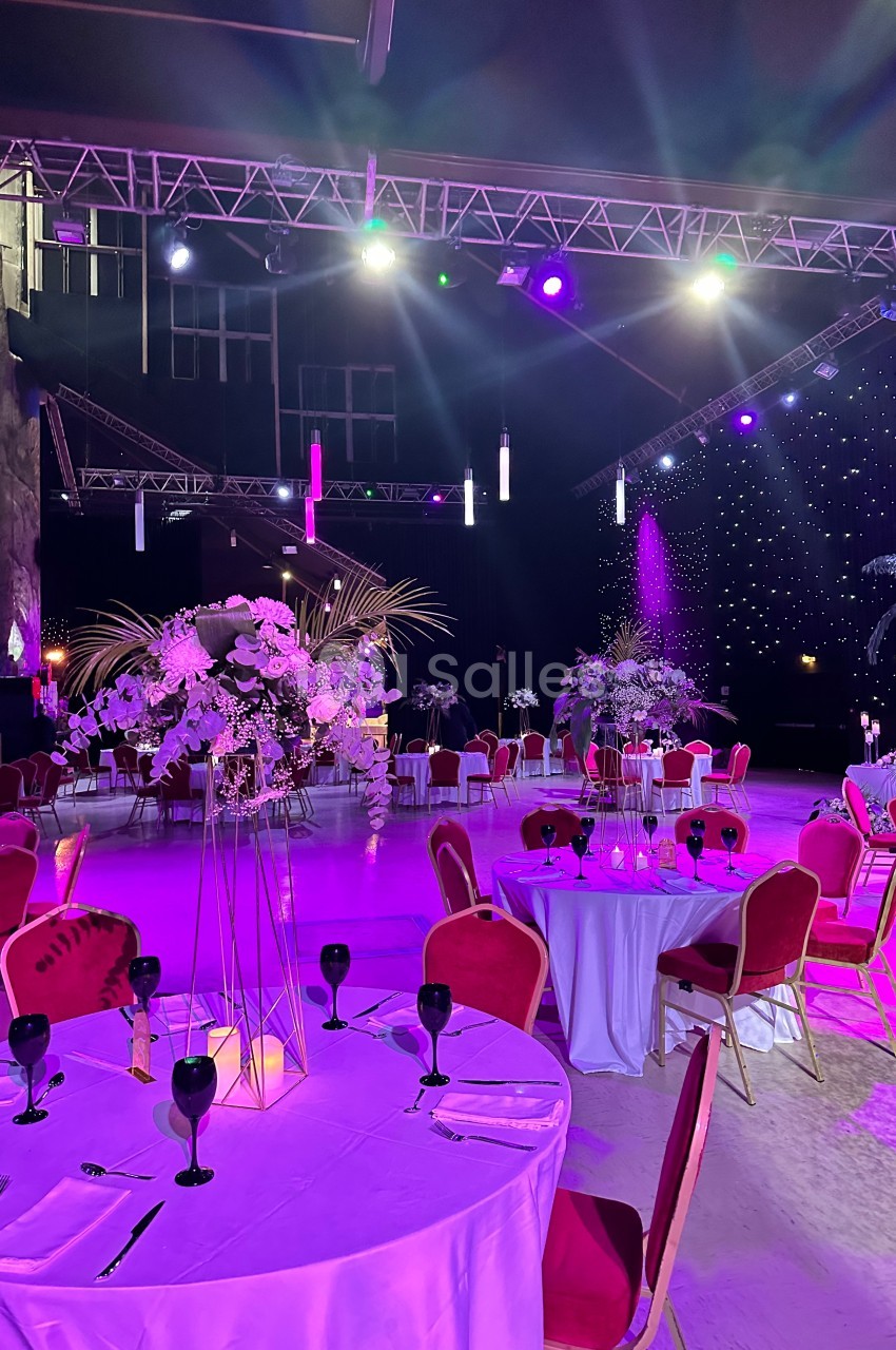 Salle de réception décorée avec des tables rondes, nappes blanches, chaises rouges et éclairage violet.