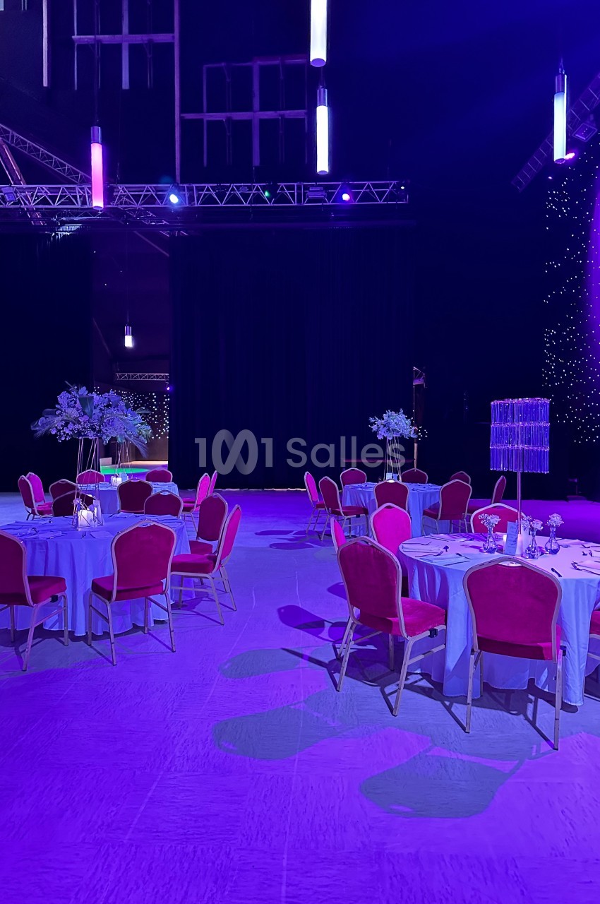 Salle de réception décorée avec des tables rondes, nappes blanches, chaises rouges et éclairage violet.