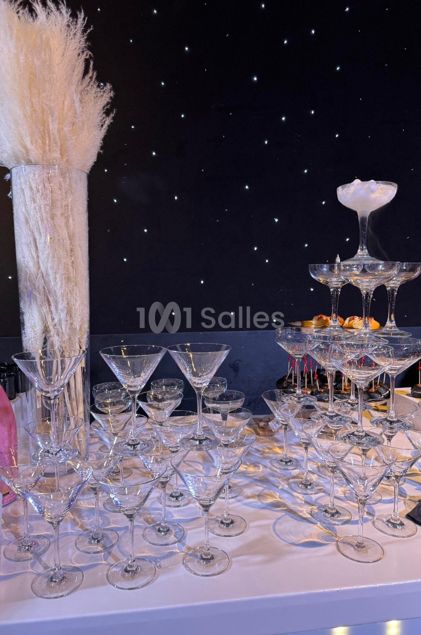 Table décorée avec des verres à cocktail empilés en pyramide, un vase de pampas et une bouteille rose, sur fond étoilé.