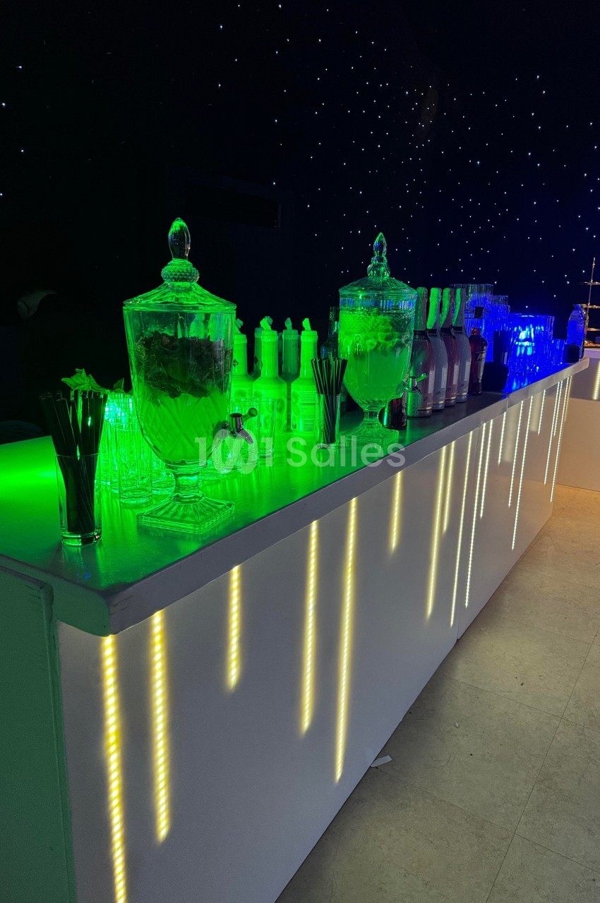 Bar éclairé avec des LED colorées, contenant des jarres de boissons, des verres et des accessoires de service.