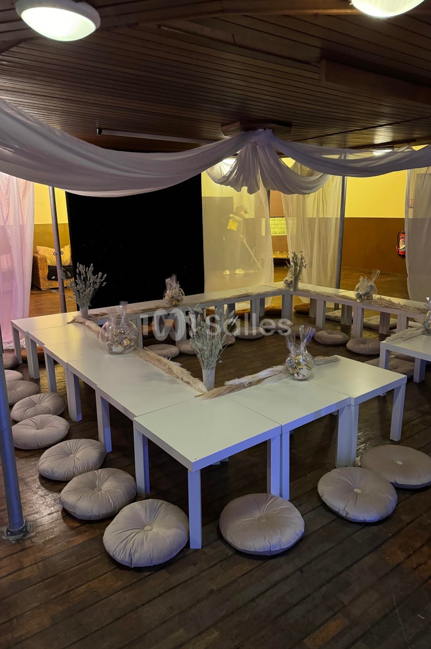 Tables blanches disposées en U avec des coussins au sol, décorées de vases et de fleurs, dans une salle éclairée.
