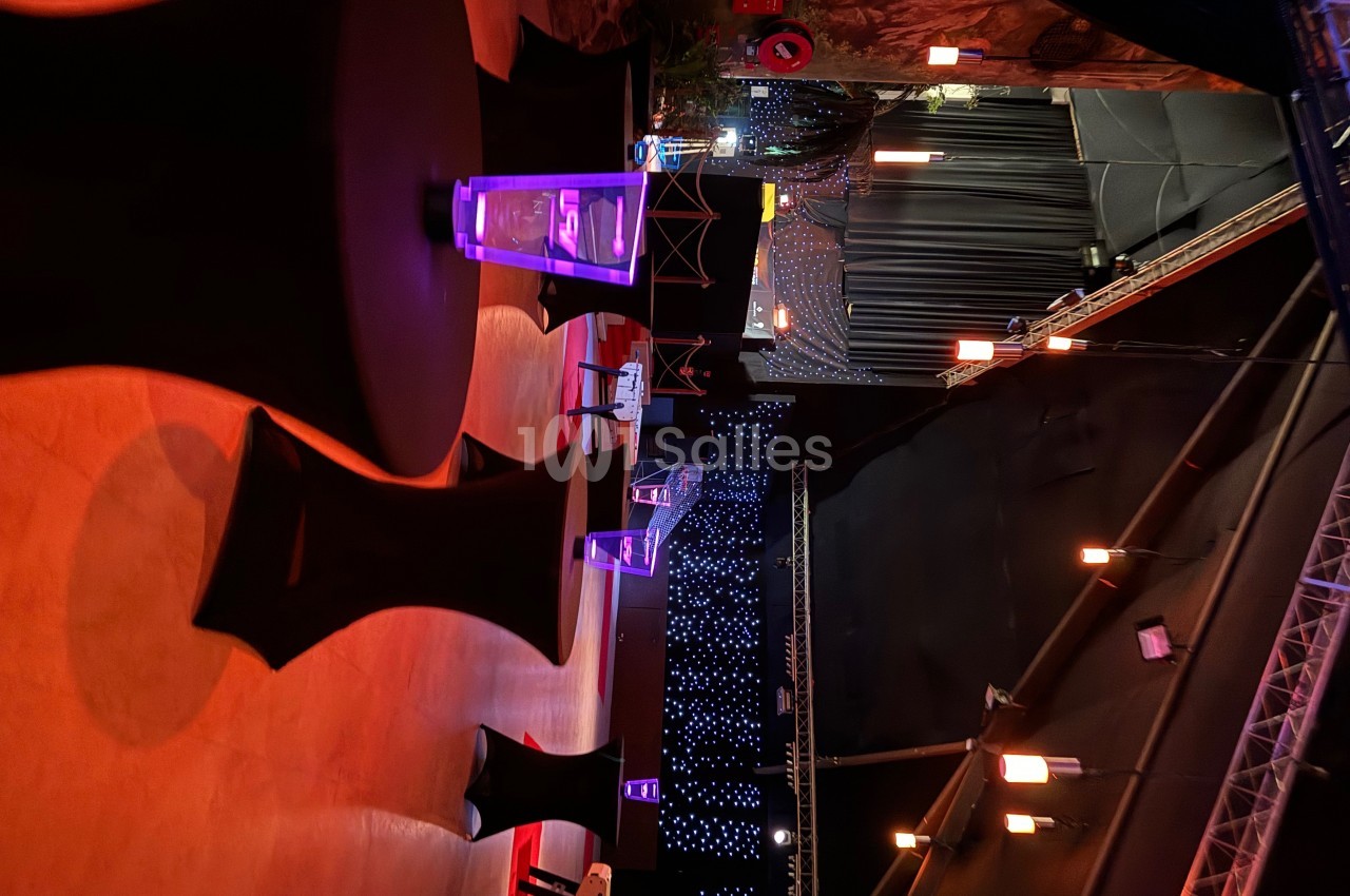 Salle sombre avec tables hautes noires, éclairage tamisé et décorations lumineuses en arrière-plan.