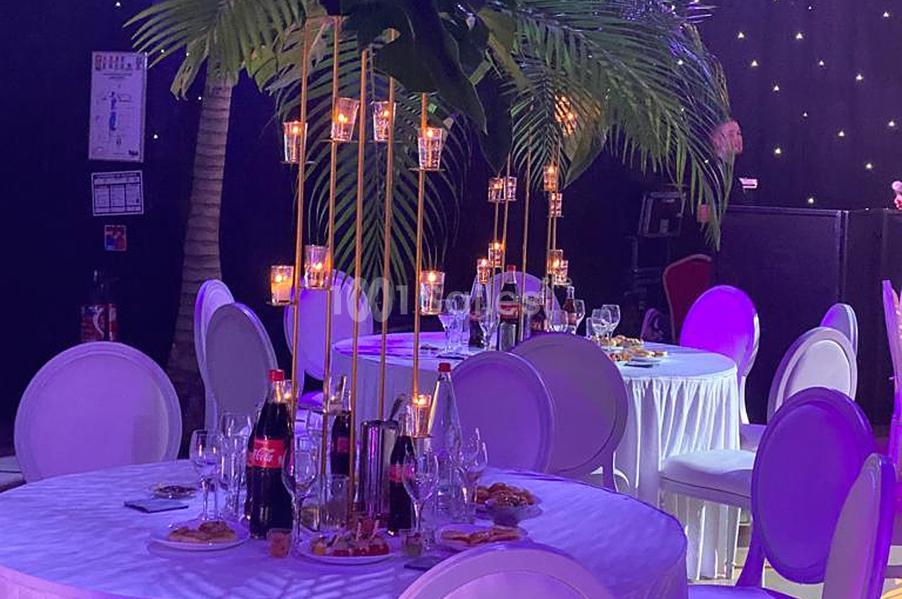 Tables rondes décorées avec des bougies suspendues, des bouteilles et des verres, sous des palmiers artificiels dans une…