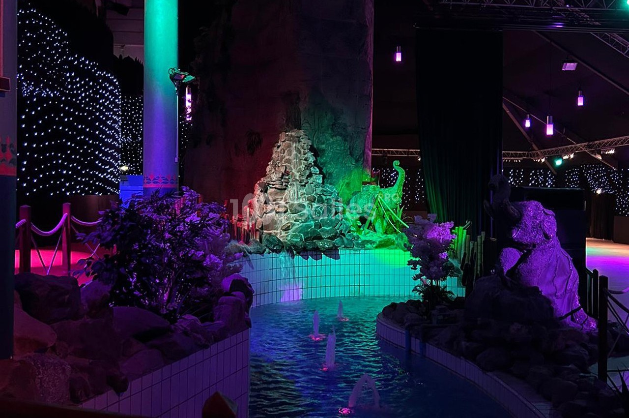 Décor intérieur avec bassin éclairé, rochers, statues et lumières colorées dans une ambiance tamisée.