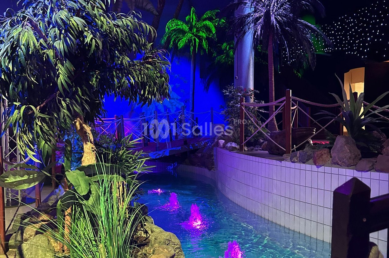 Piscine intérieure décorée de plantes tropicales, éclairée par des lumières colorées et entourée d'un pont en bois.