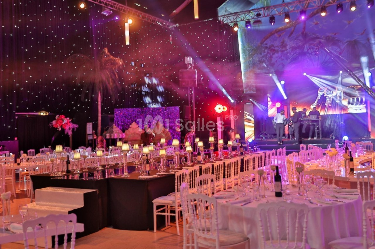 Salle décorée pour un événement avec tables dressées, éclairages colorés et scène en arrière-plan.