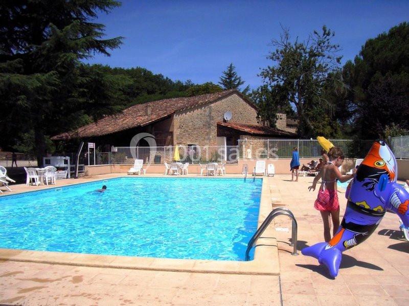 Piscine extérieure avec des transats autour, des enfants jouant et une maison en pierre en arrière-plan.