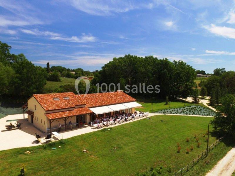 Bâtiment avec toit en tuiles entouré de verdure, terrasse ombragée et invités assis, ciel dégagé en arrière-plan.