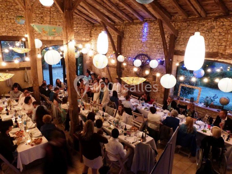 Salle en pierre avec des convives assis à des tables décorées, éclairée par des guirlandes lumineuses et des lanternes…