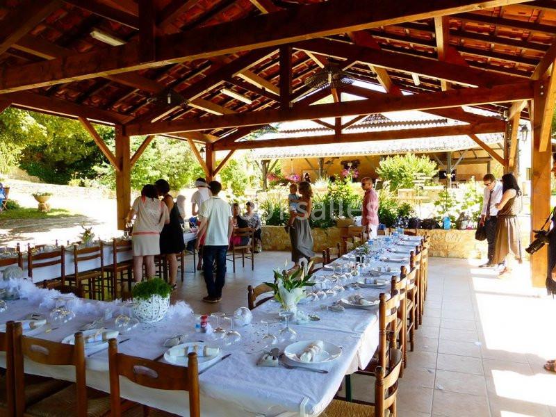 Tables dressées pour un repas sous une pergola en bois, avec des invités discutant en arrière-plan dans un cadre champêtre.