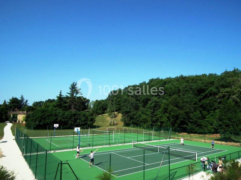 Terrain de tennis extérieur entouré de verdure, avec plusieurs joueurs en action sous un ciel dégagé.