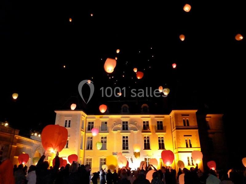 Des lanternes lumineuses s'élèvent dans le ciel nocturne devant un grand bâtiment éclairé, entouré de spectateurs.