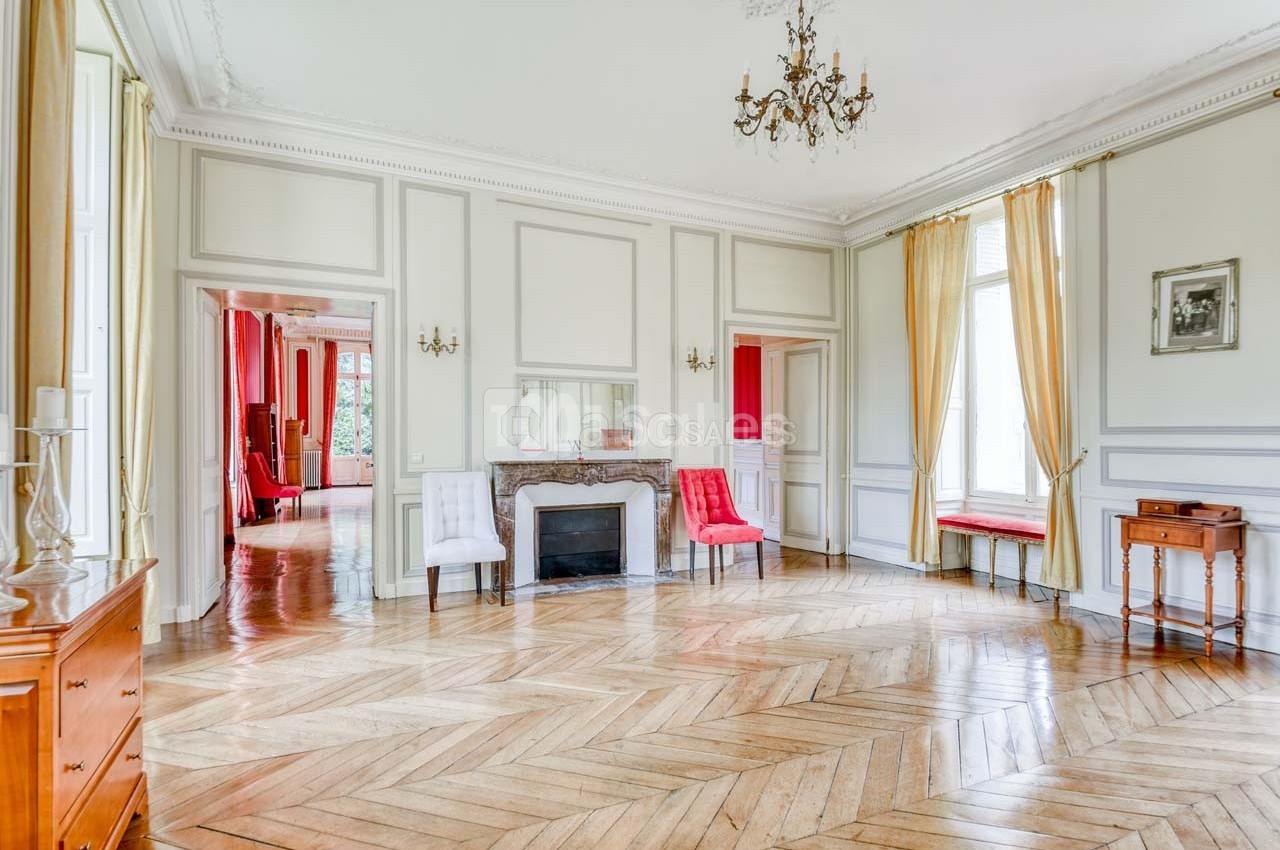 Salon lumineux avec parquet en bois, cheminée décorative, chaises et grandes fenêtres encadrées de rideaux jaunes.