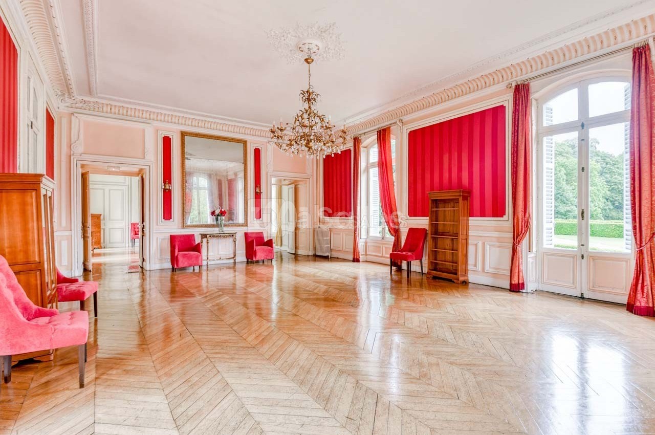 Salle lumineuse avec parquet en bois, murs rouges, moulures blanches, lustre doré et grandes fenêtres donnant sur un jardin.