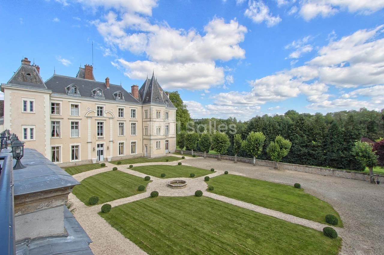 Façade d'un château historique entouré d'une cour avec pelouse, buis taillés et allées gravillonnées, sous un ciel…