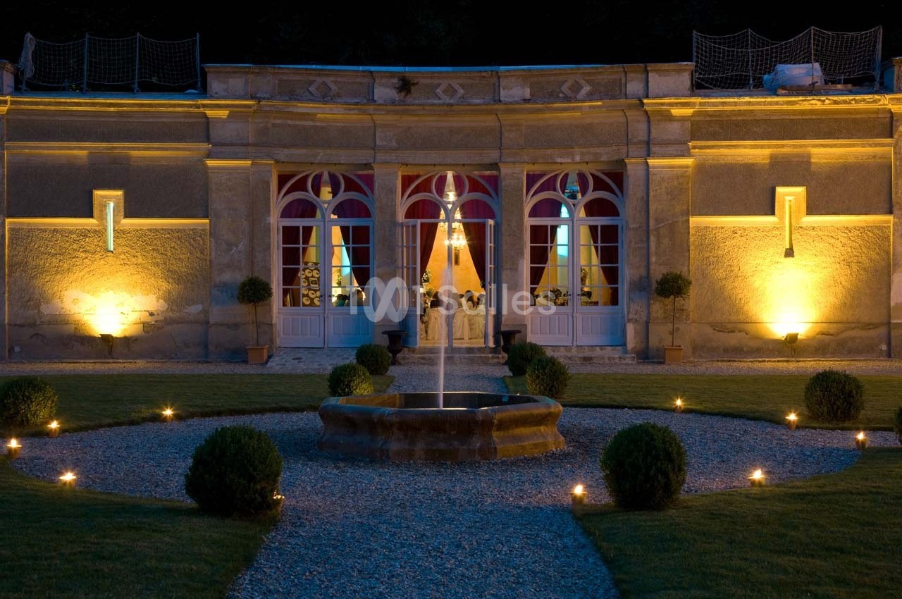 Façade d'un bâtiment éclairé la nuit avec une fontaine centrale entourée de pelouse et de buissons.