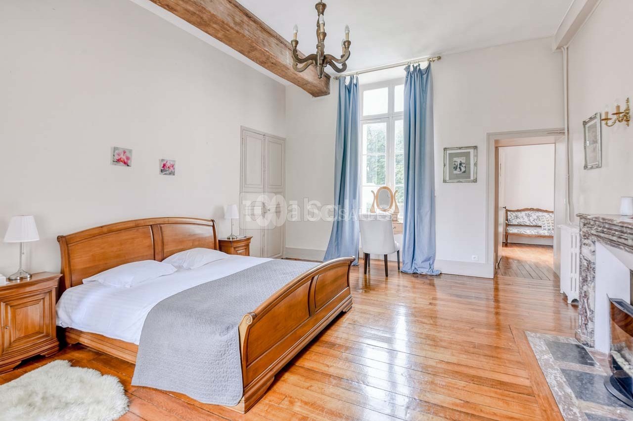 Chambre lumineuse avec lit double en bois, parquet, cheminée, poutre apparente et rideaux bleus devant une grande fenêtre.