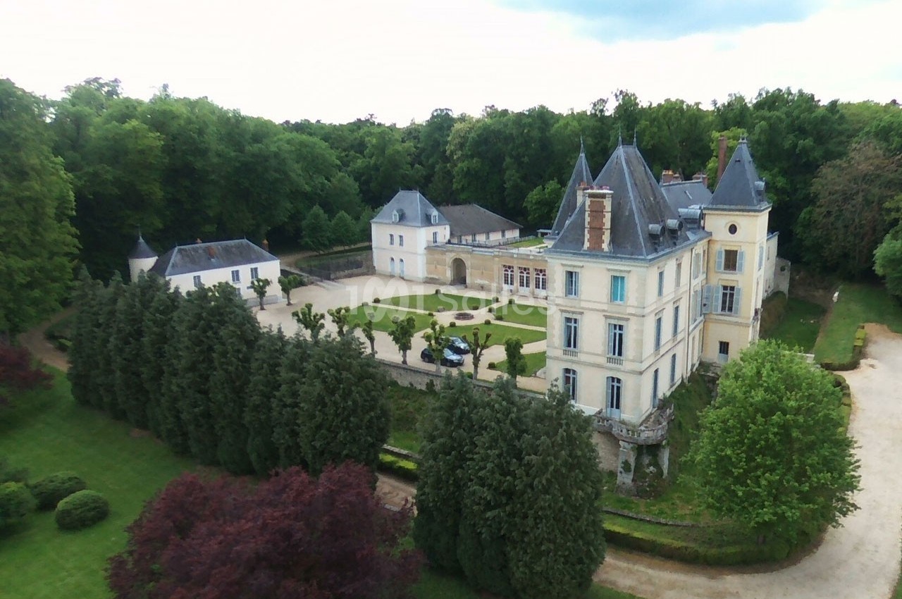 Vue aérienne d'un château entouré de jardins, d'arbres et d'une cour centrale avec une voiture stationnée.