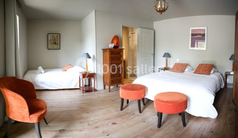 Chambre lumineuse avec deux lits, mobilier en bois, fauteuils et tabourets orange, et une salle de bain attenante.