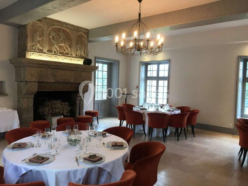 Salle lumineuse avec tables rondes dressées, chaises rouges, cheminée en pierre sculptée et lustre suspendu.
