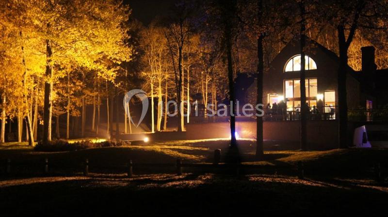 Maison éclairée la nuit entourée d'arbres aux feuilles dorées, avec un jardin sombre au premier plan.