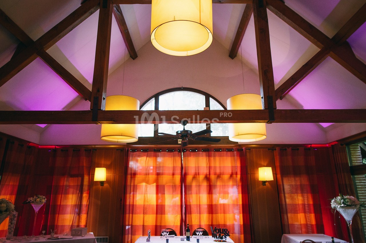 Salle avec poutres apparentes, rideaux rouges et éclairage chaleureux, décorée pour un événement avec des tables dressées.