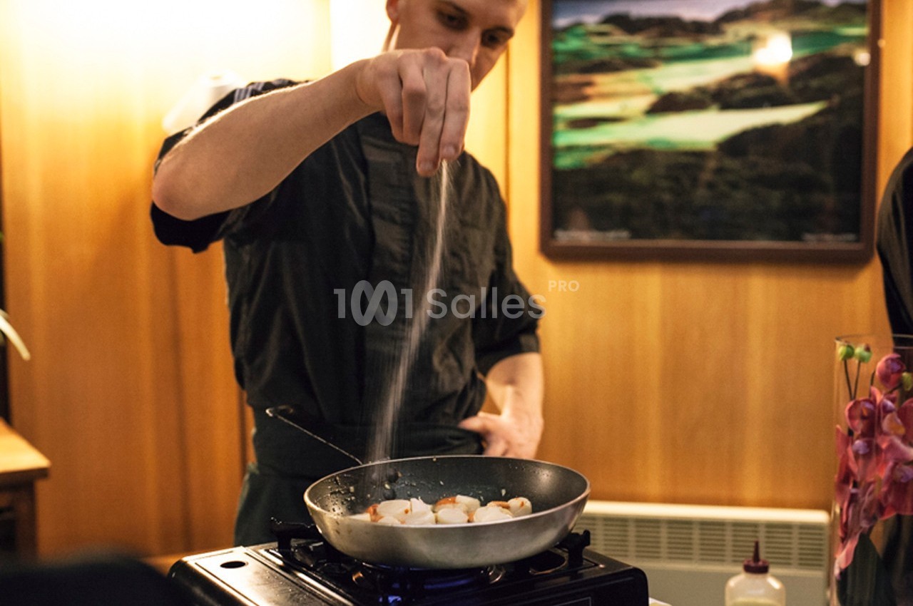 Un cuisinier saupoudre du sel sur des coquilles Saint-Jacques en train de cuire dans une poêle sur un réchaud.