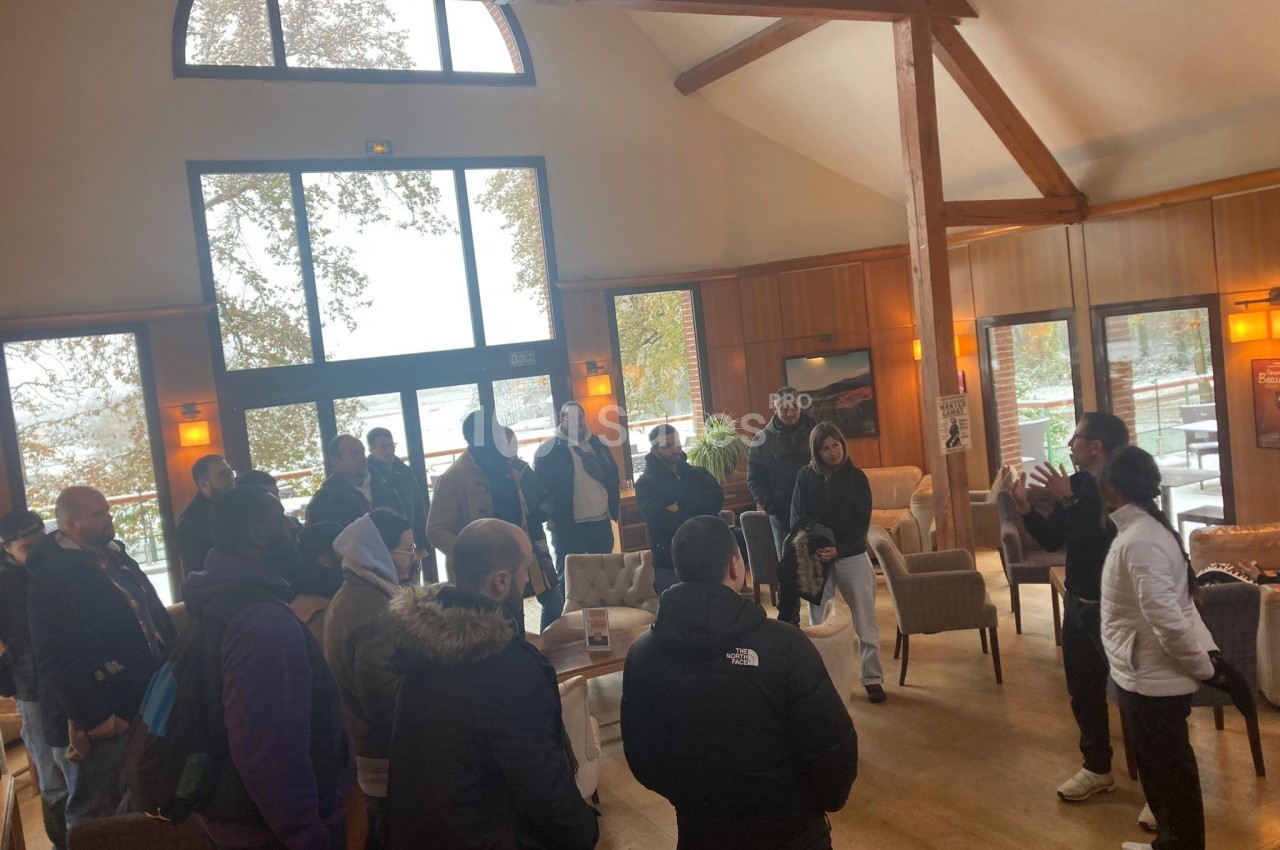 Un groupe de personnes debout dans une salle lumineuse avec de grandes fenêtres et un plafond en poutres apparentes.