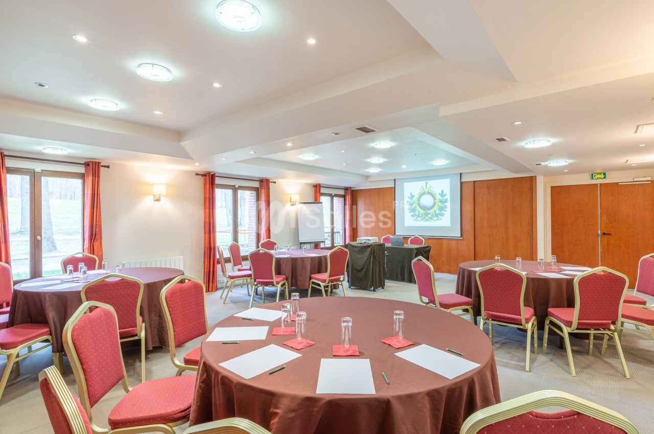 Salle de réunion lumineuse avec tables rondes, chaises rouges, écran de projection et fenêtres donnant sur l'extérieur.