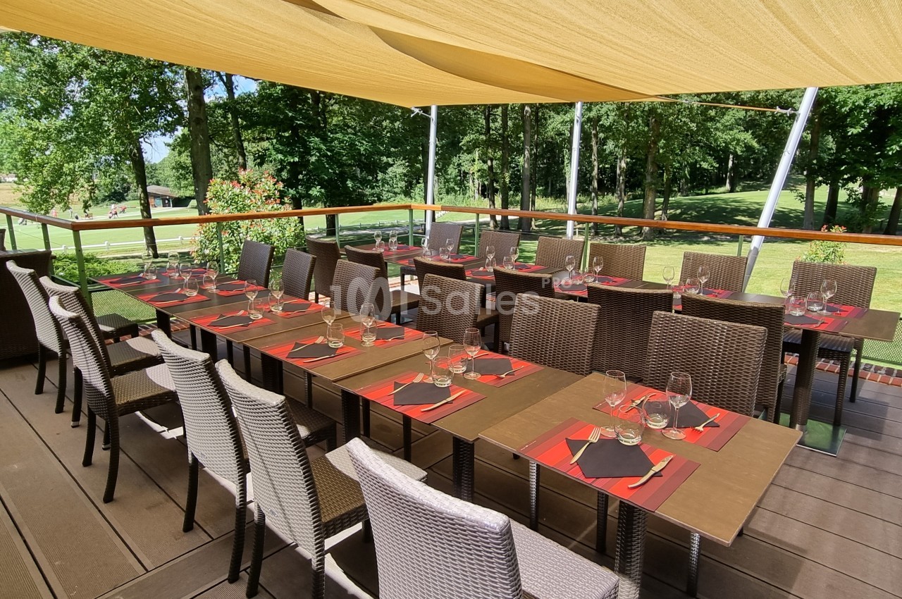 Terrasse ombragée avec tables dressées, chaises en osier et vue sur un espace vert arboré.