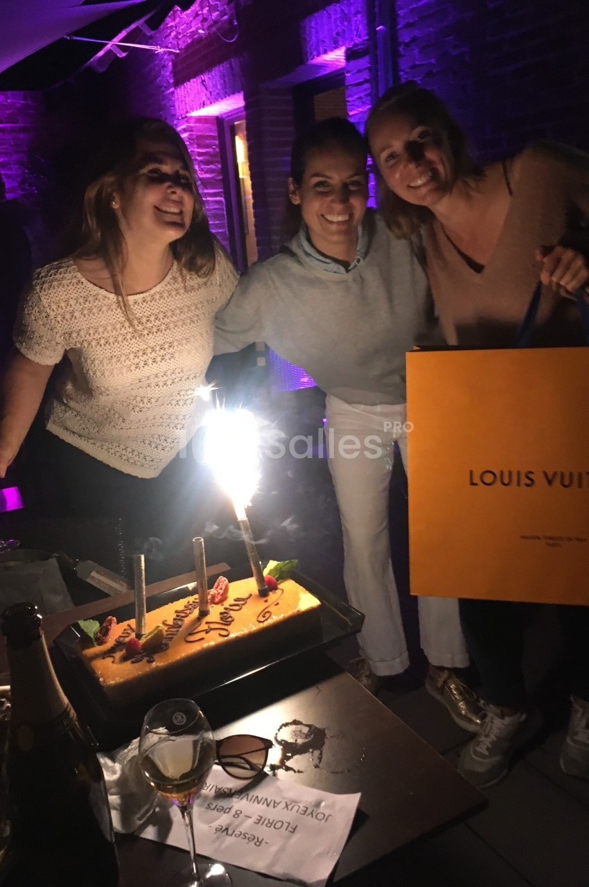 Trois femmes souriantes autour d'un gâteau d'anniversaire éclairé par une bougie étincelante, dans un cadre nocturne.