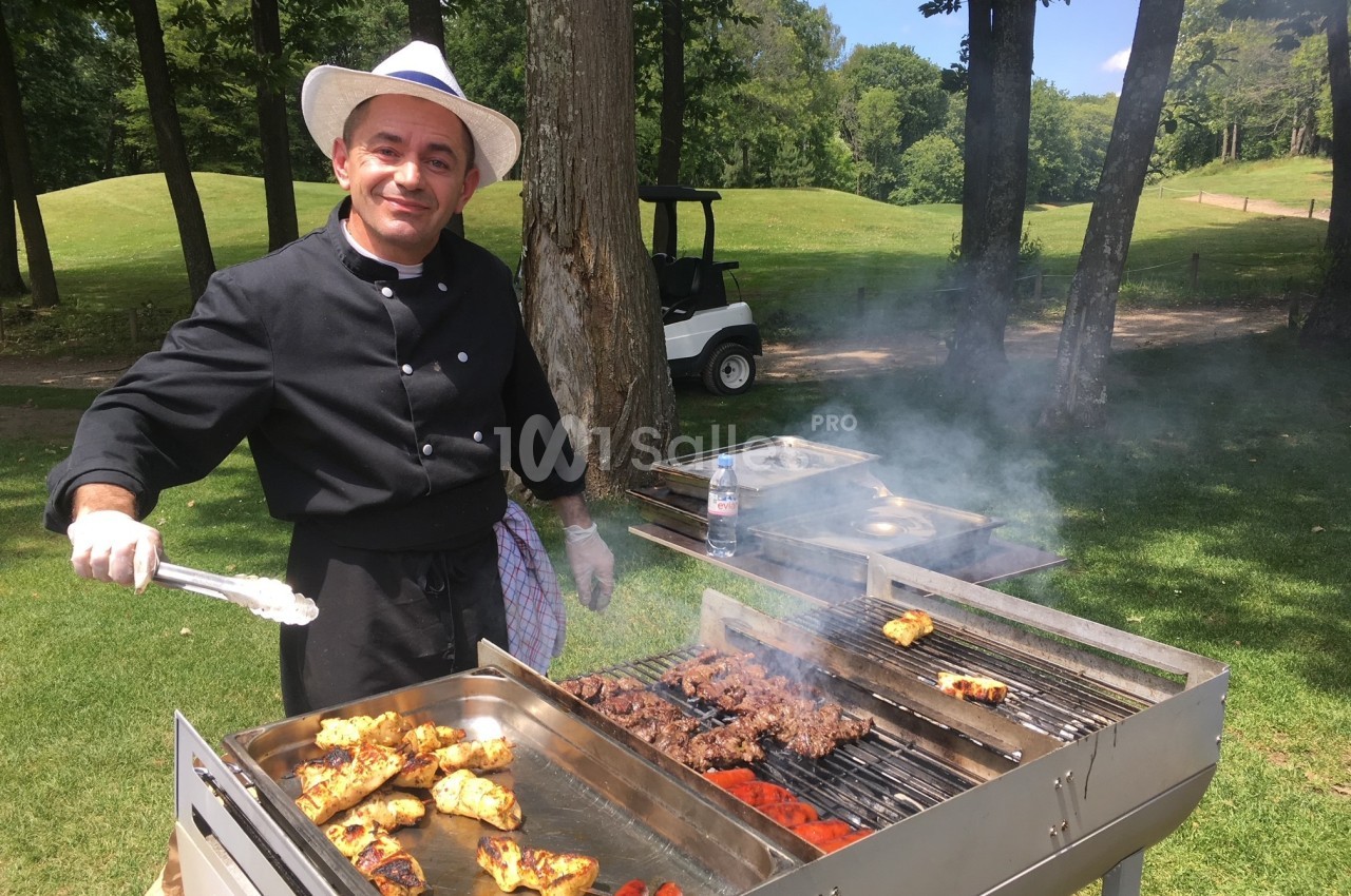 Un cuisinier en tenue noire prépare des grillades variées sur un barbecue en plein air, près d'un terrain verdoyant.