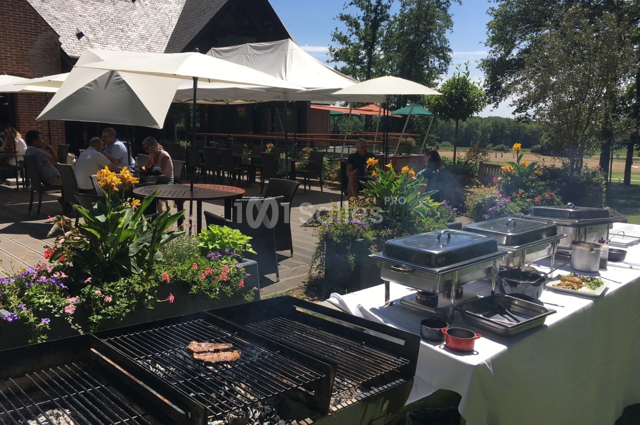 Barbecue en plein air sur une terrasse fleurie avec des tables ombragées et des convives en arrière-plan.