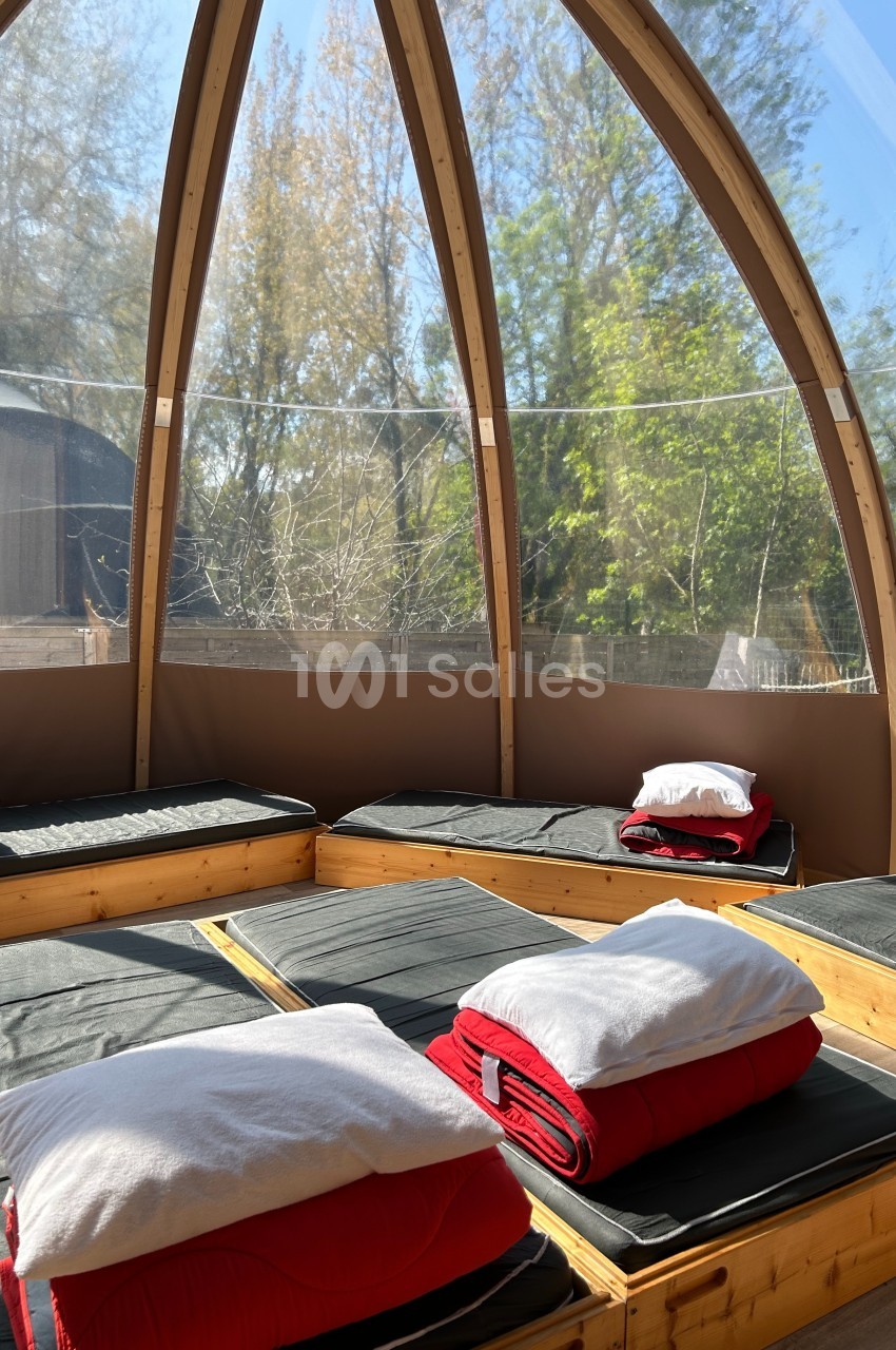 Intérieur d'une structure en bois avec des matelas et des couvertures rouges, entourée de grandes baies vitrées donnant sur…