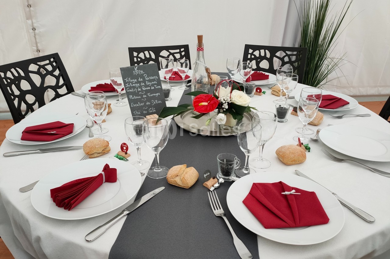 Table dressée pour un repas, avec nappes blanches et grises, serviettes rouges pliées, vaisselle, pain et décoration florale.