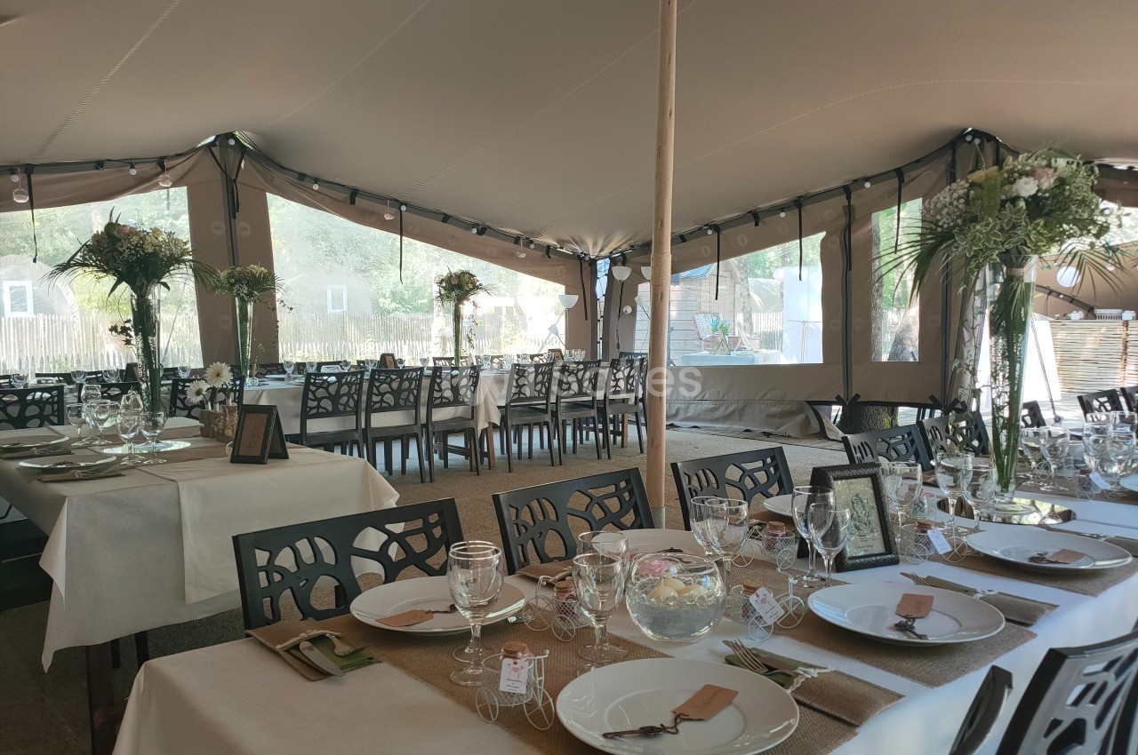Salle de réception sous une tente avec tables décorées de nappes blanches, vaisselle élégante et arrangements floraux.
