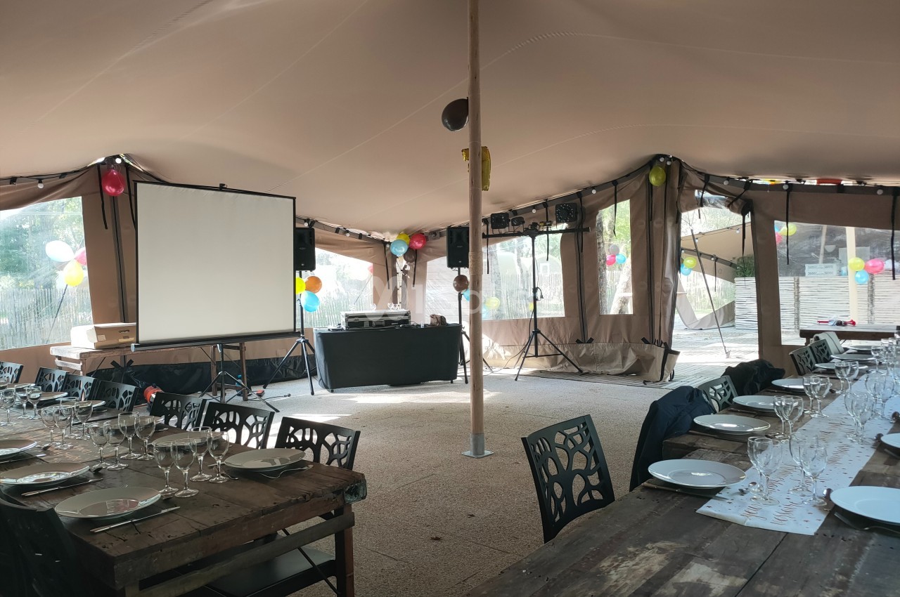 Salle de réception sous tente avec tables dressées, écran de projection, décorations colorées et équipement audio.