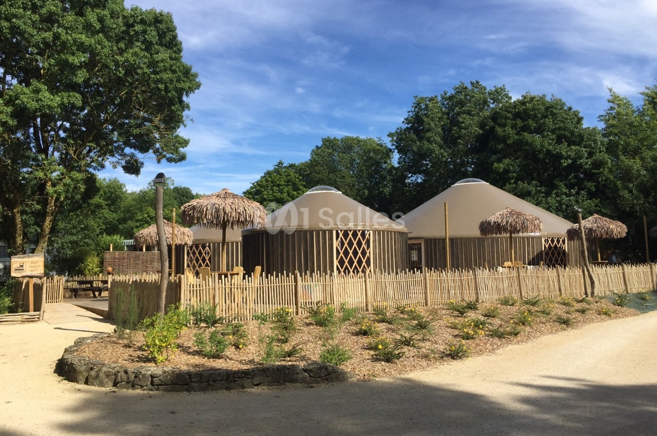 Ensemble de yourtes en bois entourées de palissades, situées dans un environnement arboré par une journée ensoleillée.