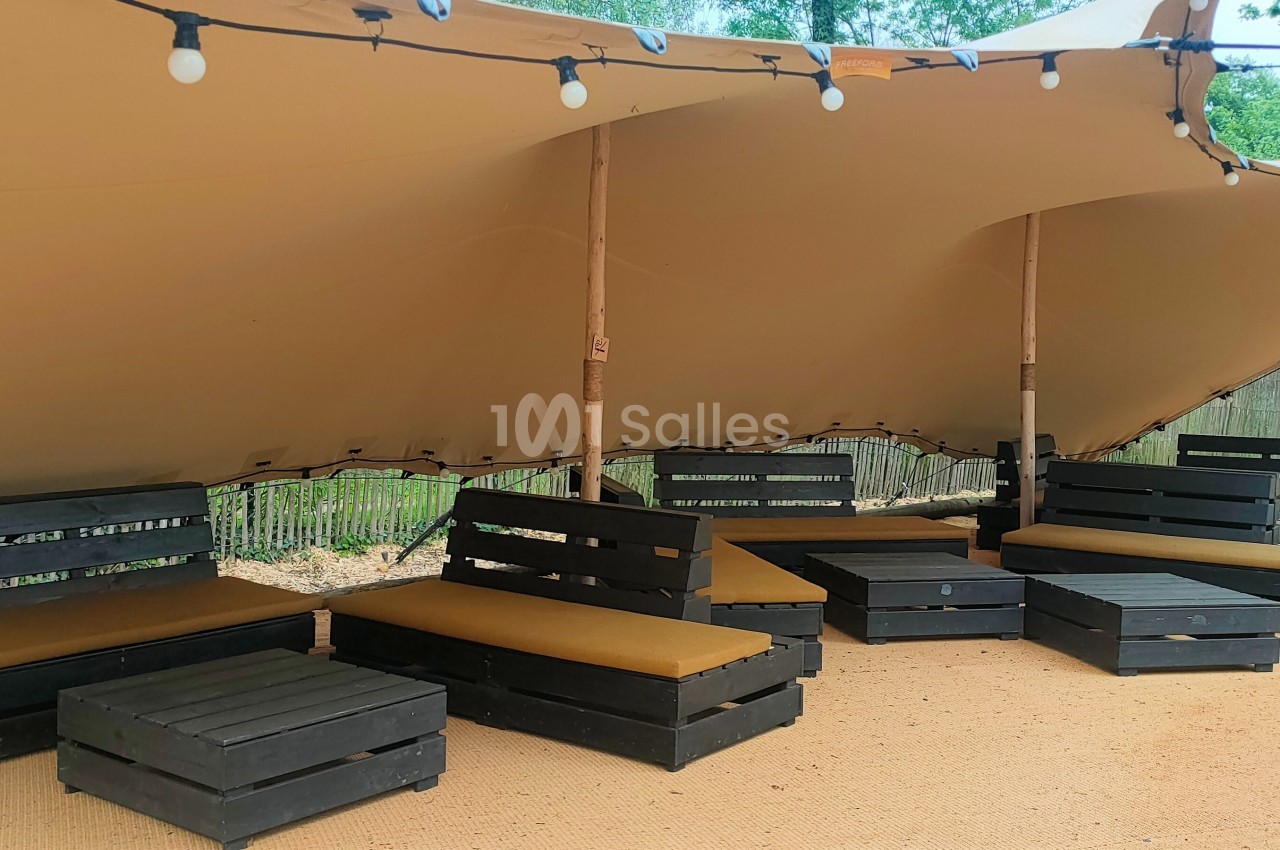Mobilier en palettes avec coussins sous une toile tendue, éclairé par des guirlandes lumineuses, dans un espace extérieur.