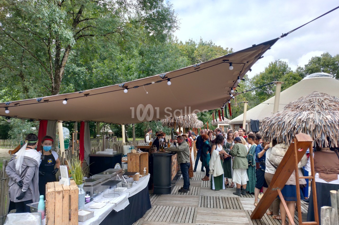 File d'attente devant un stand de restauration en plein air, sous une grande toile tendue, entouré d'arbres.