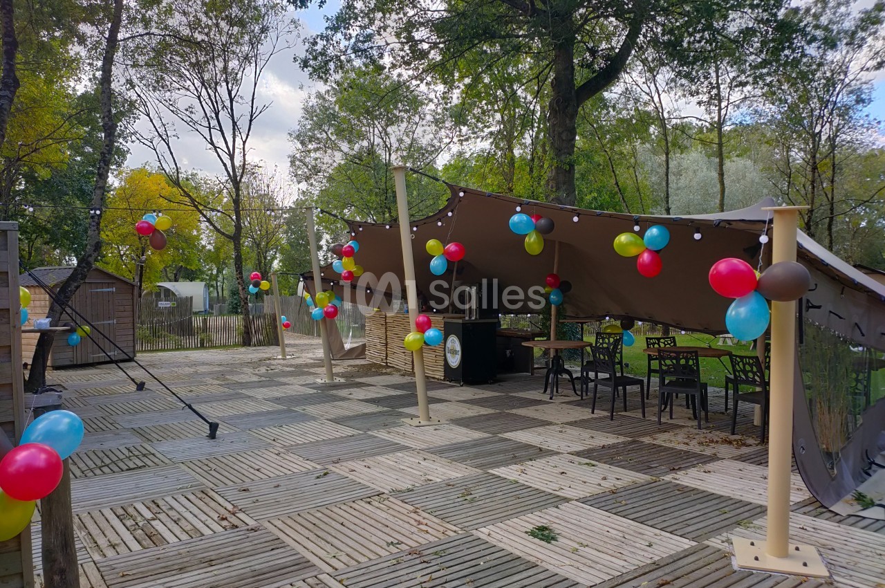 Terrasse en bois décorée de ballons colorés sous une grande tente, entourée d'arbres en arrière-plan.