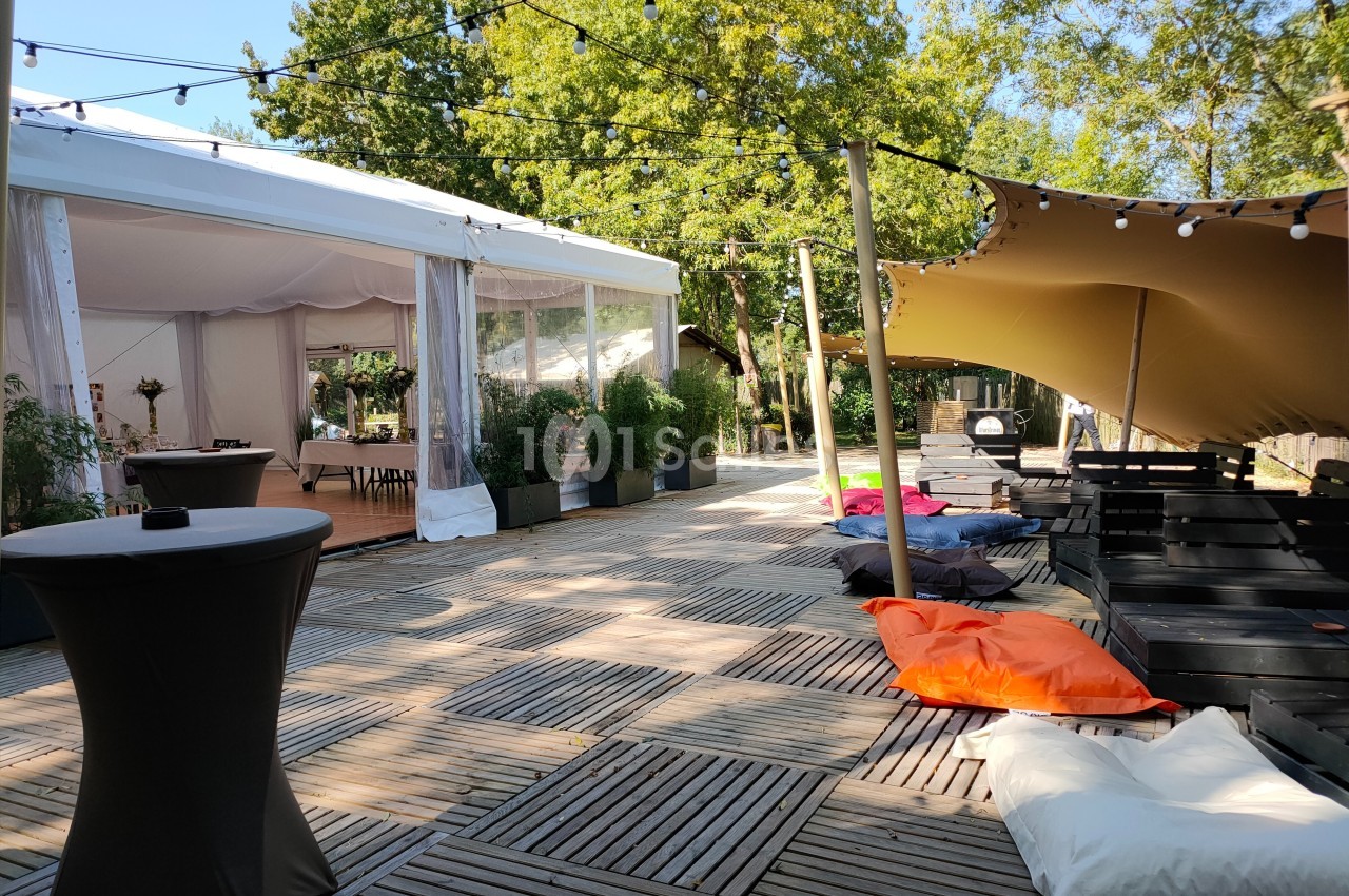 Terrasse en bois avec des coussins colorés, une tente blanche, des guirlandes lumineuses et des arbres en arrière-plan.